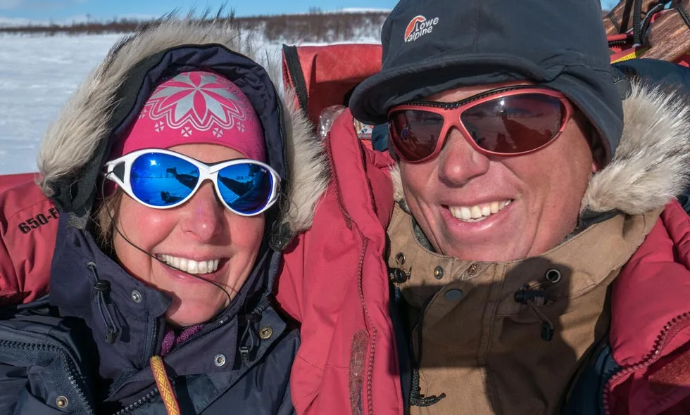 Ett par i vinterkläder med solglasögon och mössor ler mot kameran i ett kallt, snöigt landskap