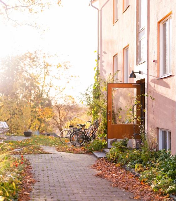 Solig höstdag vid bostadshus med öppen dörr, cyklar och löv längs stenlagd gång.