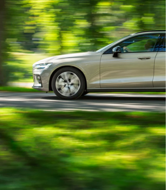 En silverfärgad kombibil kör på en slingrig landsväg omgiven av lummiga gröna träd och skog, vilket ger en känsla av en lugn bilresa i naturen