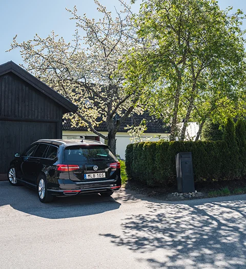 Svart bil parkerad vid en uppfart framför ett hus, omgiven av grönska och träd i blom.