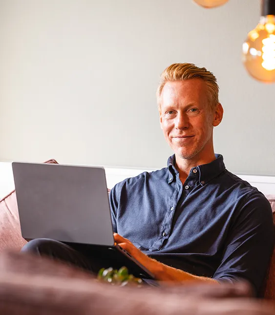 En man med kort blont hår sitter på en soffa och ler medan han arbetar på sin laptop