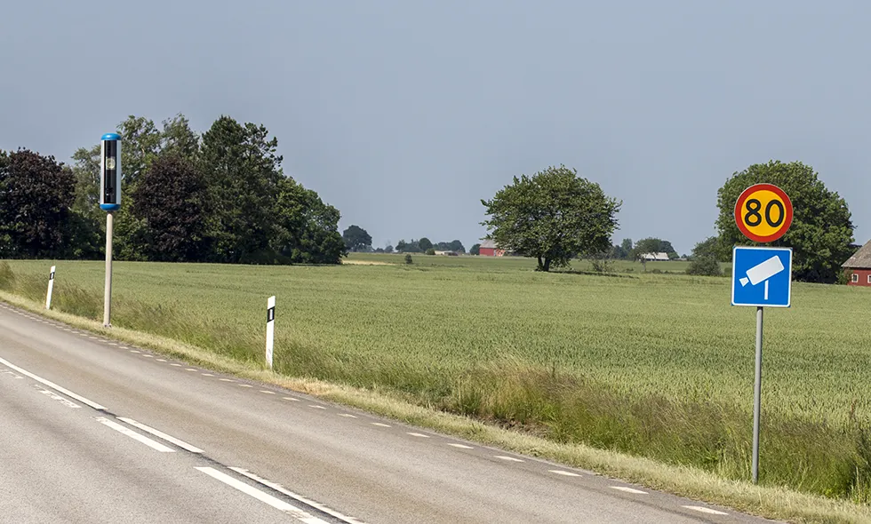 Hastighetsskylt för 80 km/h och varningsskylt för fartkamera samt fartkamera längs en landsväg omgiven av åkrar och träd.