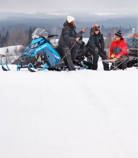 Fyra personer sitter på snö med snöskotrar, grillar och umgås i vinterlandskap.