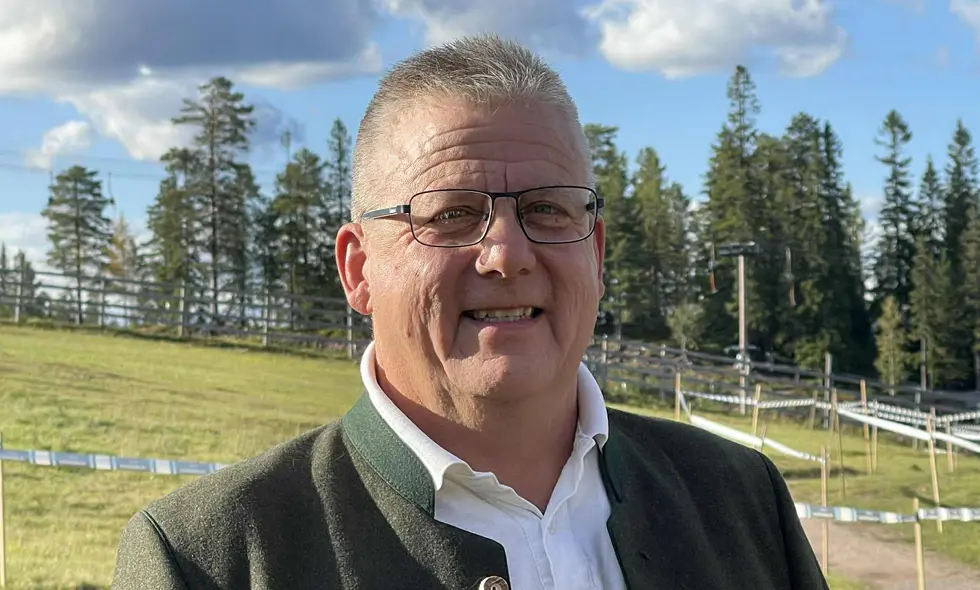 Leende man med glasögon och grått hår poserar utomhus i traditionell jacka, med grön sluttning och tallar i bakgrunden.