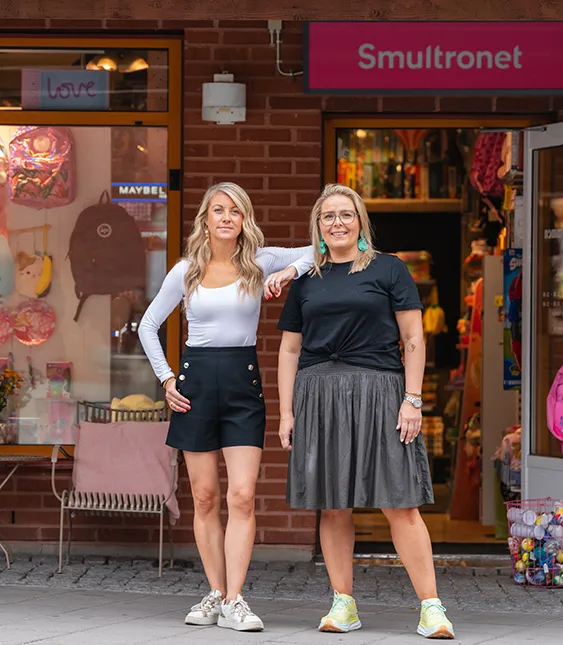 Två glada kvinnor står utanför en butik med fönsterskyltning med leksaker och accessoarer i rosa och vita färger.