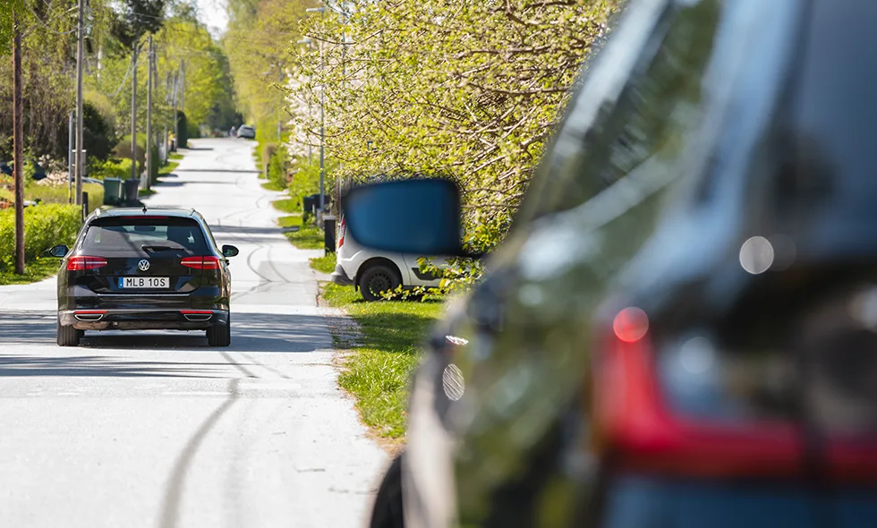 Bil kör på en solig villagata omgiven av gröna träd och parkerade bilar.