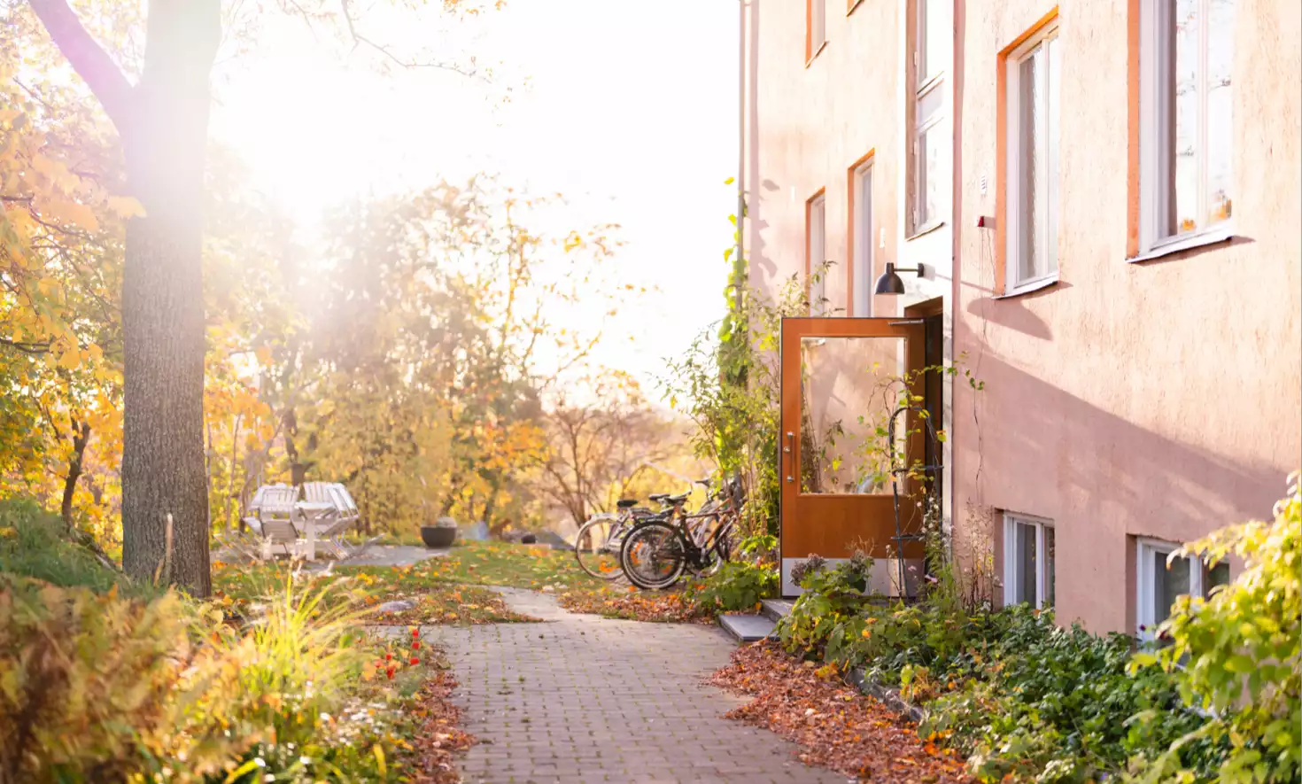 Solig höstdag vid bostadshus med öppen dörr, cyklar och löv längs stenlagd gång.