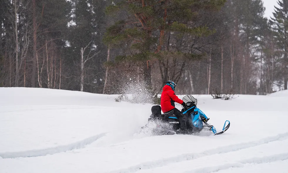 Person på snöskoter i vinterlandskap, klädd i röd jacka och blå hjälm, kör genom snö med träd i bakgrunden och snö som yr upp.