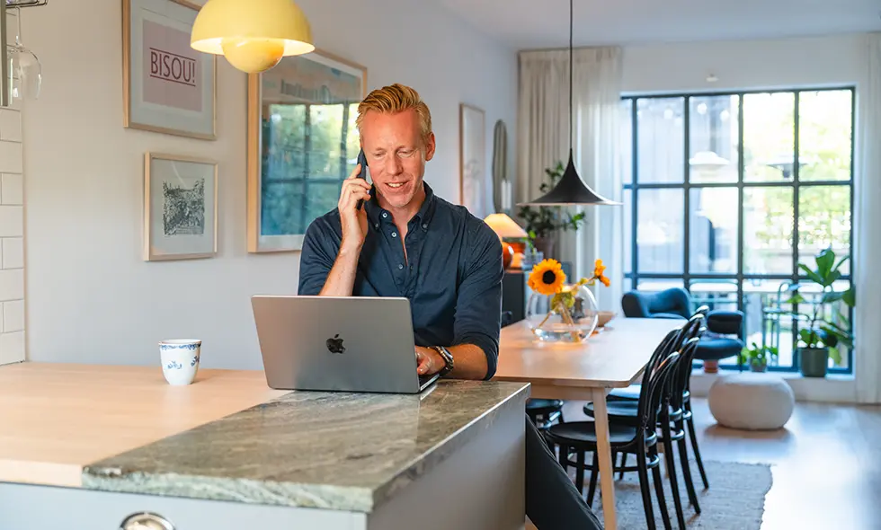 Man arbetar på en bärbar dator och pratar i telefon vid ett köksbord i ett modernt hem med ljus och växter.