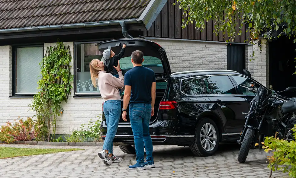 Familj vid uppfart som packar bilens bagageutrymme bredvid en vit villa och en motorcykel.