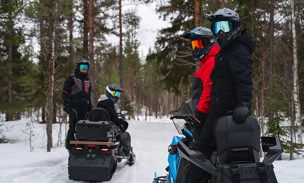 Fyra personer på snöskotrar i en snötäckt skog, alla iförda hjälmar och vinterkläder, njuter av en paus i vinternaturen.