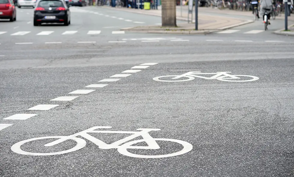 Cykelfält med vitmålade cykelsymboler i en stadskorsning. Bilar och en cyklist syns i bakgrunden på vägen.