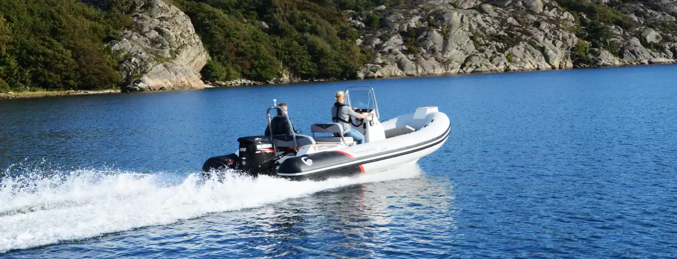Ribbåt med utombordsmotor av märket Suzuki körs av två personer på en sjö med klippor och skog i bakgrunden.