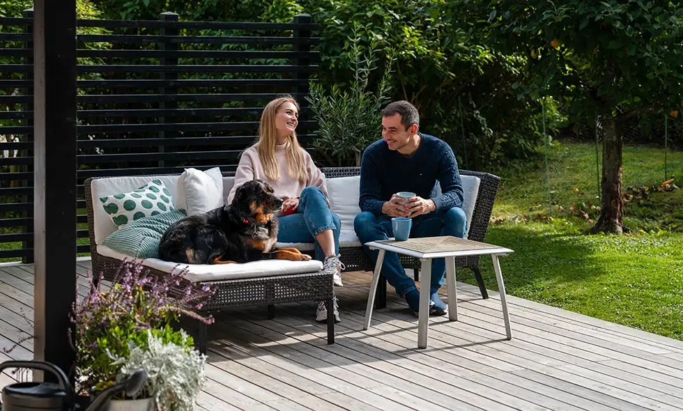 En man och en kvinna sitter och pratar på en altan med en hund liggande bredvid, omgivna av grönska.