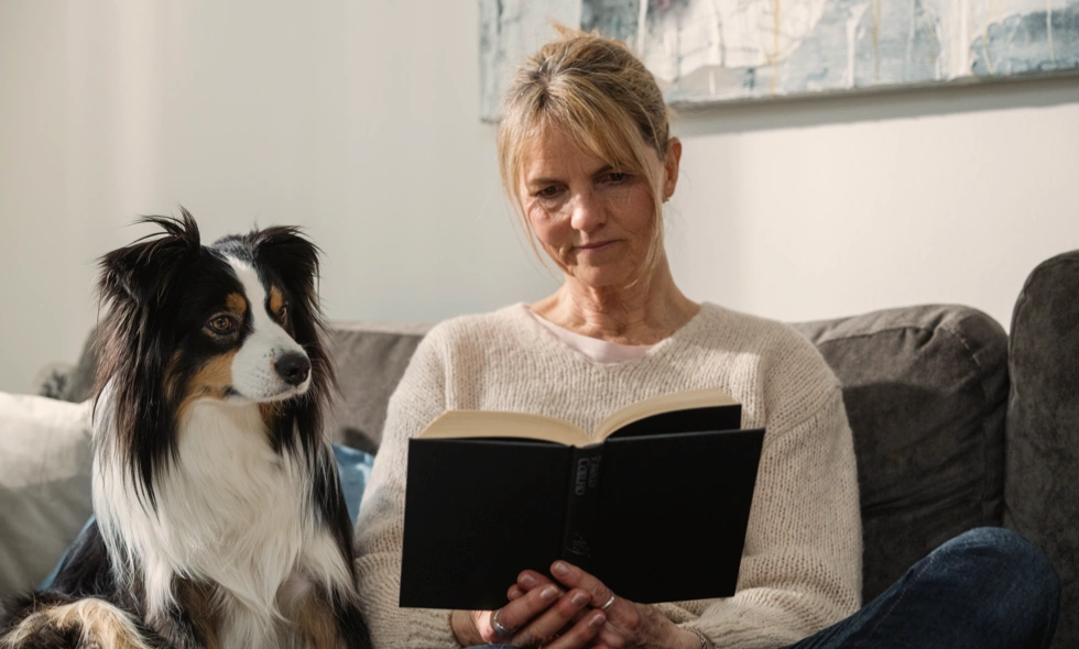 Kvinna sitter i en soffa och läser en bok med en hund bredvid sig.