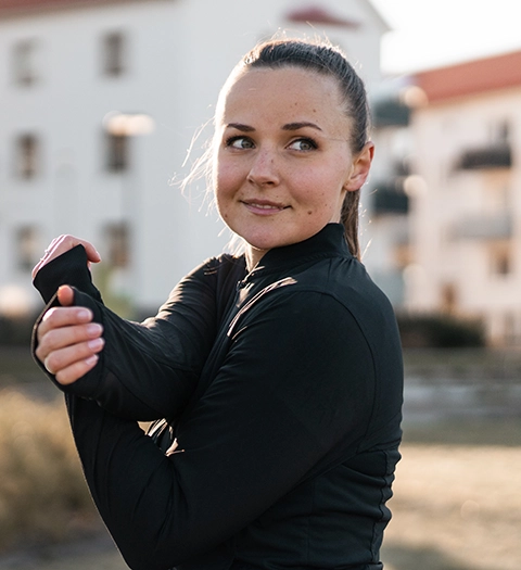 Kvinna i träningskläder som stretchar armarna, med suddig bakgrund av bostadshus.