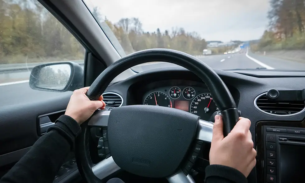 Förarens händer håller i ratten på en bil som färdas på motorvägen. Vägen sträcker sig framåt med skog och trafik i bakgrunden.