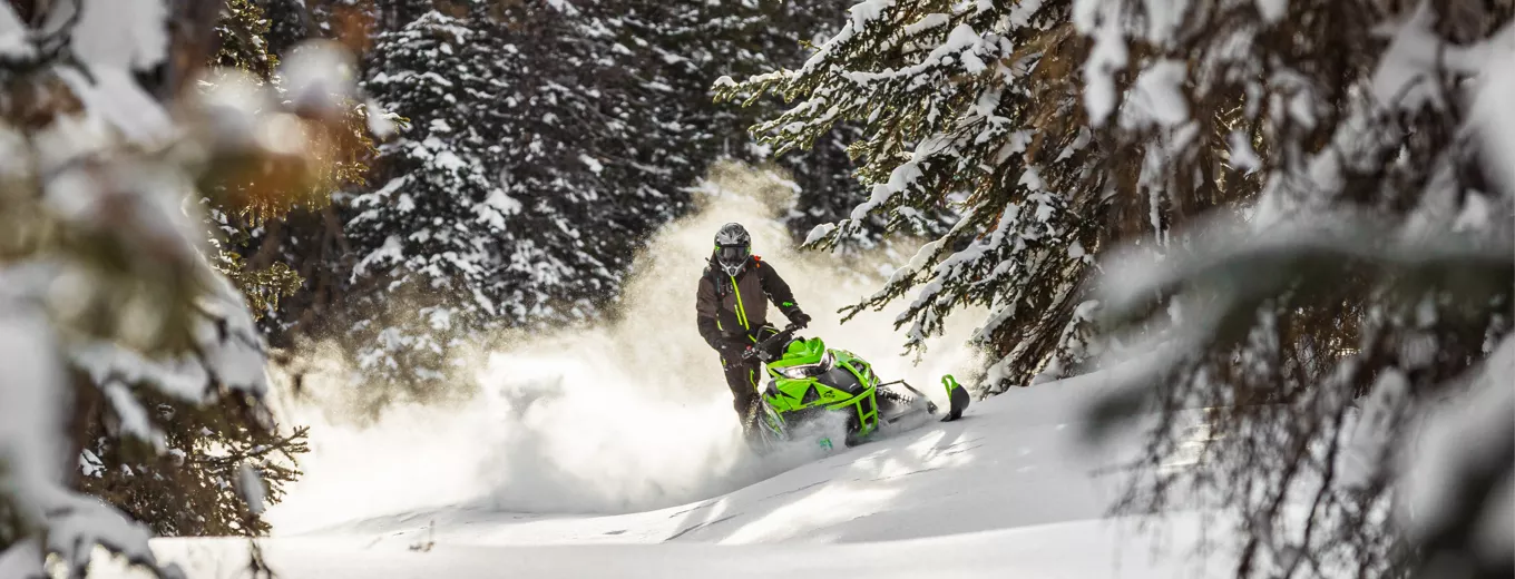 Person kör grön snöskoter av märket Arctic Cat genom snöig skog med granar.