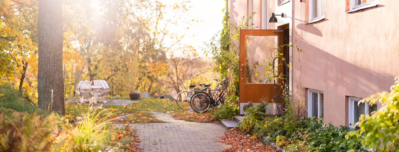 Solig höstdag vid bostadshus med öppen dörr, cyklar och löv längs stenlagd gång.