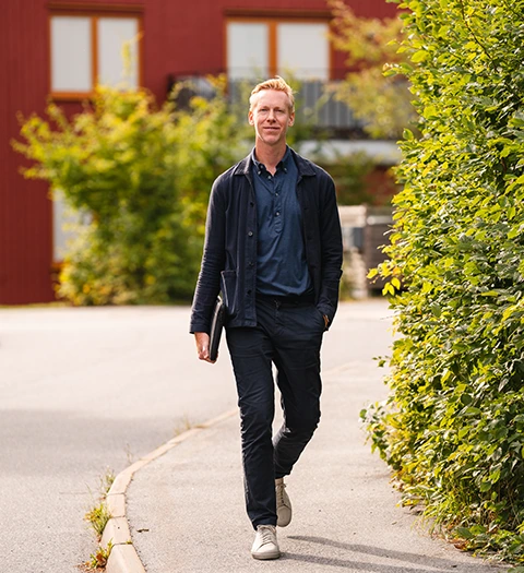 Man går på en trottoar omgiven av grönska, med en byggnad i bakgrunden, och en laptop i fodral i handen.