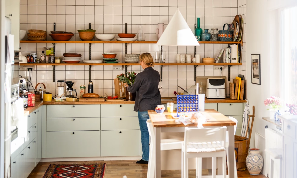 Ljust kök med öppna hyllor, kaklade väggar och en kvinna som ordnar med blommor vid bänken.