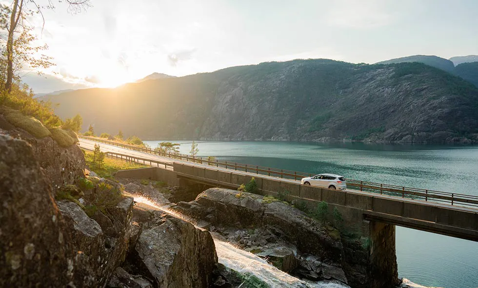 Bil kör över bro vid solnedgång, omgiven av berg och sjö.