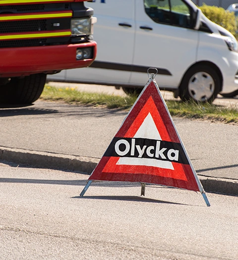 Skylt med texten "Olycka" placerad vid vägkanten, med räddningsfordon och skåpbil i bakgrunden.