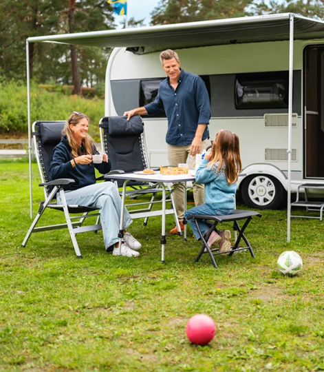 Familj fikar utomhus vid husvagn, med campingmöbler och fotbollar på gräsmattan.