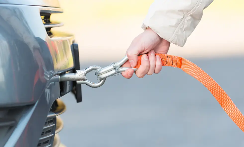 En person fäster en orange bogserlina i en bils bogserögla. Närbild av handen och metallkopplingen på fordonets front.