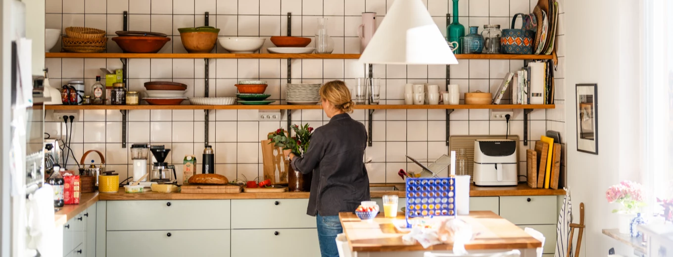 Ljust kök med öppna hyllor, kaklade väggar och en kvinna som ordnar med blommor vid bänken.