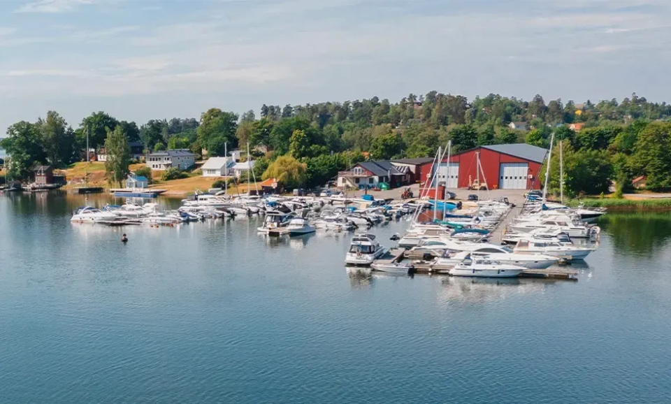 Båtar i en båtklubbshamn omgivna av vatten, på mark är det grönskande träd.