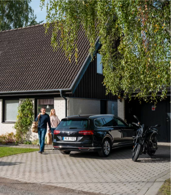 Villa med vitt tegel, mörkt tak och stenlagd uppfart. Ett par lämnar huset och går mot en parkerad bil. Bredvid bilen står en mc.