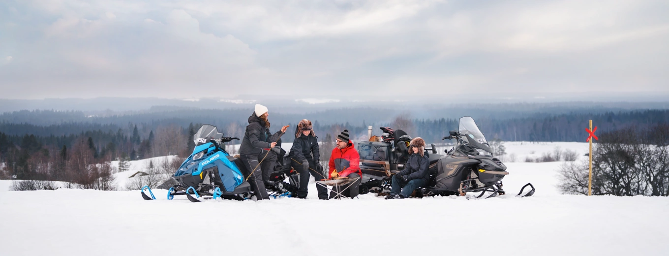 Fyra personer sitter på snö med snöskotrar, grillar och umgås i vinterlandskap.